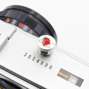 以下が含まれることがあります： 黒いダイヤルと、赤と白のピンポンラケットのデザインが施されたボタンが付いた、銀色のカメラ本体のクローズアップ。3054800の数字が本体に印刷されています。オレンジ色のインジケーターが見えます。