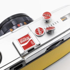 May include: Silver vintage camera with yellow leather trim, featuring red Coca-Cola logo accents. The camera has a black lens and several Coca-Cola branded pins. The text "Enjoy Coca-Cola" is visible on the camera.