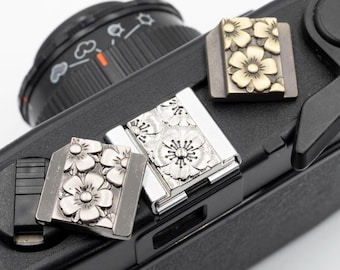 May include: Close-up of three camera hot shoe covers with floral designs. Two covers are silver-toned, and one is gold-toned. The covers are rectangular with a raised floral pattern. The covers are resting on a black camera.
