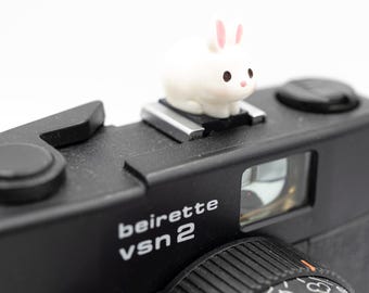 May include: A black Beirette VSN 2 camera with a small white bunny figurine with pink ears and cheeks on top. The camera has a lens and a viewfinder. The text "beirette VSN 2" is printed on the camera body.