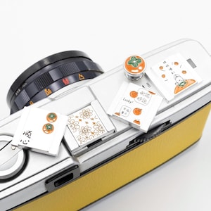 以下が含まれることがあります： 花柄、漫画のイラスト、漢字など、さまざまなデザインの小さな装飾用マグネットのコレクション。 マグネットは、シルバーとイエローのカメラ本体に配置されています。