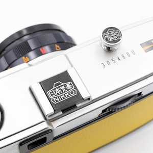 以下が含まれることがあります： 黄色の革製グリップが付いた、ヴィンテージの銀色のカメラのクローズアップ。カメラには黒いレンズ、"NIPPON KOGAKU TOKYO"の文字が入った銀色のボタン、日本語のテキストと"NIKKO"の文字が入った黒い長方形のパーツがあります。