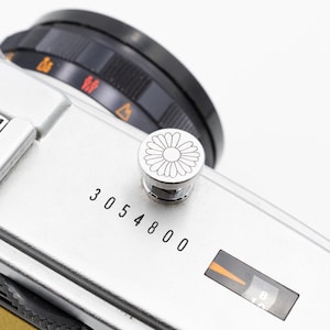 以下が含まれることがあります： 黒いレンズダイヤルと花の模様の銀色のボタンが付いた、銀色のカメラ本体のクローズアップ。 シリアル番号3054800がカメラ本体に印刷されています。 カメラにはオレンジと黒のダイヤルが付いています。