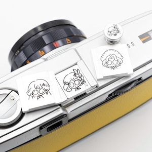 以下が含まれることがあります： 黒いレンズと黄色の革製グリップが付いた銀色のカメラ。漫画のキャラクターのイラストが描かれた3つの小さな長方形の銀色のアクセサリーがカメラに配置されています。同様のイラストが描かれた丸い銀色のアクセサリーが上にあります。