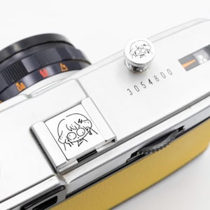 以下が含まれることがあります： 黄色の革製グリップが付いた、シルバーのヴィンテージカメラ。 カメラは黒いレンズと、それぞれ漫画のキャラクターイラストが描かれたシルバーのボタンとシルバーのプレートを備えています。 シリアル番号「3054800」がカメラ本体に印刷されています。