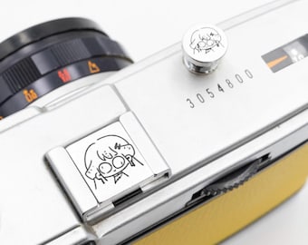 May include: A silver vintage camera with a yellow leather grip. The camera features a black lens and a silver button and a silver plate, each with a cartoon character illustration. The serial number "3054800" is printed on the camera body.