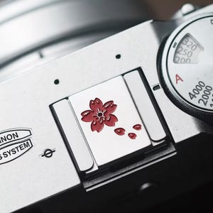 以下が含まれることがあります： 赤い桜のデザインが施された、銀色のカメラのクローズアップ。カメラには数字と文字が表示されたダイヤルが付いています。