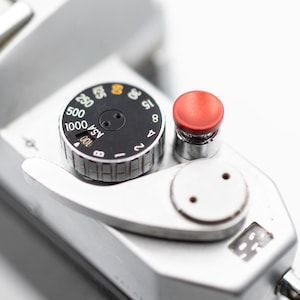 May include: Close-up of a silver camera with a black dial that has numbers from 1 to 8, 15, 20, 25, 30, 50, 100, 200, 500, 1000, and ASA. The dial also has a red button.