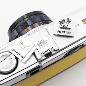 May include: A silver and yellow Fujifilm camera with three small white labels. Each label has a black illustration and the word "FUJIFILM". The camera lens is black, and the body is silver and yellow.