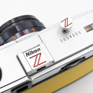 以下が含まれることがあります： 黒いレンズと黄色の革製グリップが付いた、シルバーのニコンカメラのクローズアップ。カメラには赤い「Z」ロゴと「Nikon」の文字があります。「3054800」のシリアル番号が表示されています。