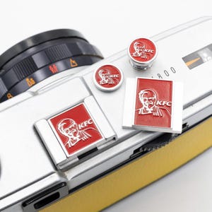 Pode incluir: Uma coleção de pinos e acessórios vermelhos e prateados com o tema KFC. Os pinos apresentam o logotipo da KFC e um retrato do Coronel Sanders. Os itens são exibidos no corpo de uma câmera prateada com uma pega de couro amarela.