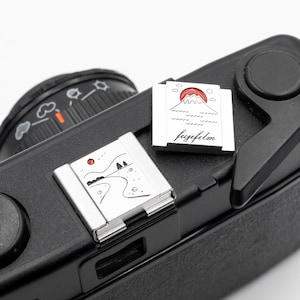 May include: Close-up of a black camera with two silver accessories. One accessory features a red sun over a mountain and the word "fujifilm". The other accessory shows a landscape with a red sun and a winding road.