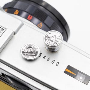 以下が含まれることがあります： 2つの装飾的なシルバーボタンが付いたシルバーカメラのクローズアップ。1つのボタンは山脈のデザインで、もう1つは日の出のある山を描いています。カメラには黒いダイヤルがあり、「54800」の数字が見えます。