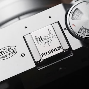 May include: Close-up of a silver Fujifilm camera. The camera features a landscape illustration with mountains, trees, and a boat. The word "FUJIFILM" is printed in bold black letters. The camera's lens system is also visible.