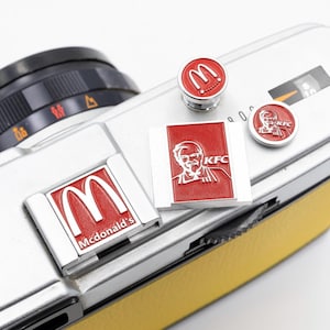 Pode incluir: Um corpo de câmera prateado com uma pega de couro amarela é adornado com vários pinos de esmalte vermelho e prateado. Os pinos apresentam os logotipos do McDonald's e KFC, incluindo os arcos dourados e o Coronel Sanders.