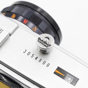 以下が含まれることがあります： 黒いレンズと山と太陽のデザインが施された装飾ボタンが付いた、ヴィンテージのシルバーカメラのクローズアップ。 カメラ本体には「3054800」の数字が印刷されています。 カメラの設定が表示されています。