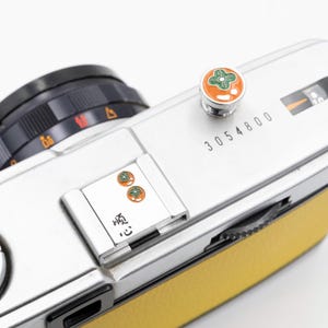 以下が含まれることがあります： 黄色い革のグリップが付いた、銀色のフィルムカメラのクローズアップ。カメラは黒いレンズ、オレンジと緑のアクセントが付いた装飾ボタン、ボディに印刷された番号「3054800」を備えています。また、ボディには「顺 心」の文字も印刷されています。