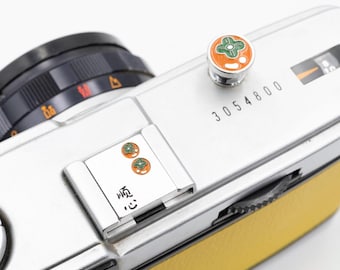 May include: A silver film camera with a yellow leather grip. The camera has a black lens, a decorative button with orange and green accents, and the number "3054800" printed on the body. The camera also has the text "顺 心" printed on the body.