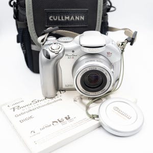 May include: Silver Canon PowerShot S1 IS digital camera with a black and gray Cullmann camera bag. The camera has a zoom lens and a lens cap. A manual and the lens cap are in the foreground.