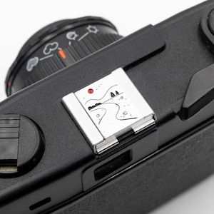 May include: Close-up of a black camera body with a silver accent. The silver accent features a small landscape illustration with a red dot. The camera lens is black with white markings.