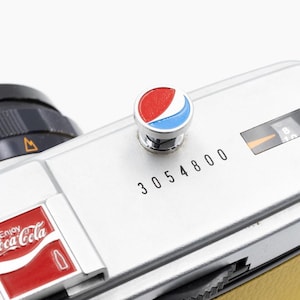 以下が含まれることがあります： ペプシのロゴボタンが付いたシルバーのカメラ本体。カメラにはコカ・コーラのロゴと数字の3054800がプリントされています。黒いレンズと黄色の革ストラップが付いています。