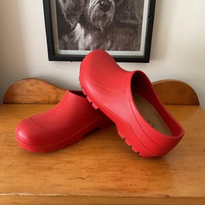 May include: A pair of bright red clogs, made of a rubber-like material, sit on a wooden surface. The clogs have a rounded toe and a low heel. A black and white framed picture of a dog hangs in the background.