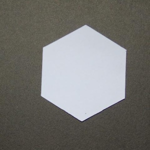 Könnte beinhalten: Eine weiße, sechseckige Form ist in der Mitte des Bildes zentriert. Die Form befindet sich auf einer dunklen, strukturierten Oberfläche. Das Hexagon hat eine einfarbige weiße Farbe.