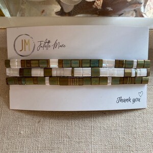 Puede incluir: Tres pulseras apiladas con azulejos blancos, verdes y dorados. Las pulseras están sobre una tarjeta blanca con el texto "Juliette Marie" y "Gracias" con un corazón.