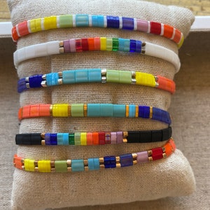 May include: A stack of colourful beaded bracelets with a rainbow pattern. The bracelets are made of small, square beads in various colours, including blue, green, yellow, orange, red, and purple. The bracelets are arranged in a stack, with the top bracelet being the most colourful.