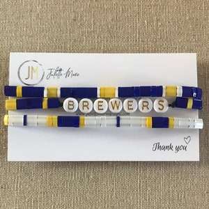May include: A white card with the text "Juliette-Marie" and "www.juliettemariedesigns.com" printed on it. The card is decorated with three beaded bracelets in blue, white, and yellow colors. The bracelets spell out the word "Brewers". The card also has the text "Thank you" with a heart symbol.
