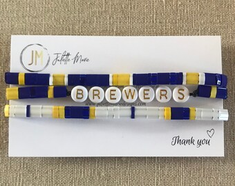Könnte beinhalten: Eine weiße Karte mit dem Text "Juliette-Marie" und "www.juliettemariedesigns.com". Die Karte ist mit drei Perlenarmbändern in den Farben Blau, Weiß und Gelb verziert. Die Armbänder buchstabieren das Wort "Brewers". Die Karte enthält auch den Text "Danke" mit einem Herzsymbol.