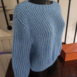 Könnte beinhalten: Hellblauer Strickpullover mit rundem Ausschnitt und langen Ärmeln. Der Pullover wird auf einer schwarzen Schaufensterpuppe präsentiert. Das Strickmuster ist sichtbar, und die Ärmel haben eine leichte Ballonform. Der Pullover ist aus weichem, strukturiertem Material.