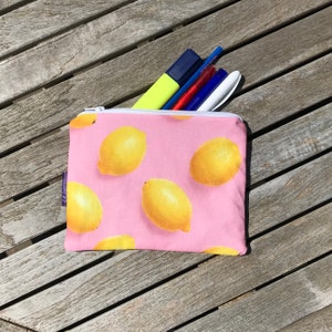 Könnte beinhalten: Ein rosa Federmäppchen mit gelben Zitronenmuster. Es enthält einen gelben Textmarker, einen blauen Textmarker, einen roten Stift, einen blauen Stift und einen weißen Stift. Auf einer Holzoberfläche.