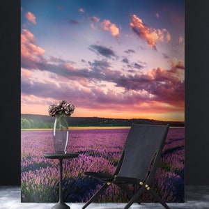 Puede incluir: Una silla negra con un marco de madera se encuentra en un campo de flores de lavanda púrpura. El cielo es una puesta de sol vibrante de color rosa y naranja con nubes esponjosas. Un jarrón de vidrio con flores moradas se encuentra en una mesa pequeña en primer plano.