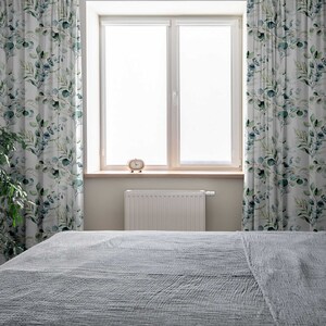 May include: A gray bed with a white radiator and a window with white curtains featuring a green leaf pattern.
