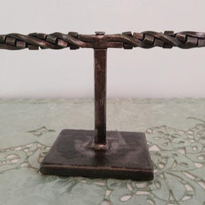 May include: A handcrafted, dark metal jewelry display stand. The stand features a square base, a vertical post, and a horizontal bar with a woven design. The metal has a rustic, aged appearance, suggesting a vintage or industrial aesthetic.