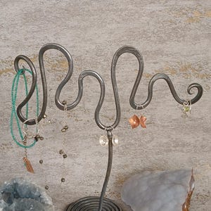 May include: A dark gray metal jewelry stand with a wavy, serpentine design featuring multiple hooks. The stand holds a green beaded necklace, a delicate silver chain with small dark beads, a pair of round silver earrings, and copper earrings.