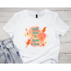 Pode incluir: Camiseta branca com a frase "Dancing Through The Lightning Strikes" em verde-azulado e laranja. O design inclui raios e estrelas laranja sobre um fundo em aquarela laranja e amarelo.