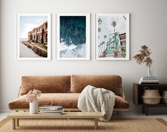 May include: Three framed photographs of a beach scene, a building with palm trees, and a wave crashing in the ocean. The photographs are hanging on a white wall above a brown couch with a white throw blanket.
