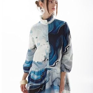 Puede incluir: Un vestido camisa con estampado azul y blanco, cuello con volantes y mangas largas. El vestido tiene un corte holgado y una falda fluida.
