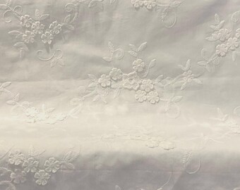 May include: White fabric with embroidered floral and leaf patterns. The embroidery features clusters of small flowers and delicate vines, creating a textured, decorative design. The fabric appears to be a light, possibly sheer material.