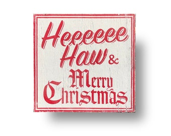 May include: Square wooden sign with a white background and red lettering. The sign reads "Heeeee Haw & Merry Christmas" in a mix of cursive and gothic fonts, framed by a red border. The wood has a distressed, rustic appearance.