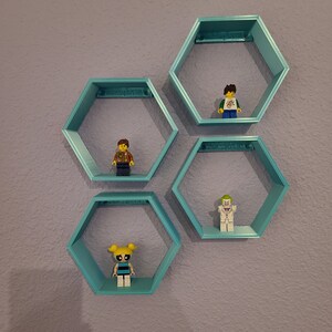 Peut inclure: Quatre étagères hexagonales turquoise montées sur un mur violet clair. Chaque étagère contient une petite figurine en plastique. L'étagère du haut a une figurine avec des cheveux bruns portant une chemise bleu et blanc. La deuxième étagère a une figurine avec des cheveux bruns portant une veste marron. La troisième étagère a une figurine avec des cheveux blancs portant un costume blanc. L'étagère du bas a une figurine avec des cheveux blonds portant une robe bleu et noir.