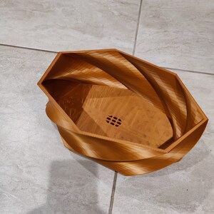 May include: A brown, wooden, geometric planter with a swirl design. The planter has a drainage hole in the bottom.