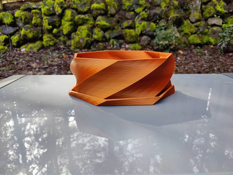 May include: A terracotta-colored, geometric planter with a swirling design. The planter is made of a smooth, textured material and has a hexagonal base.