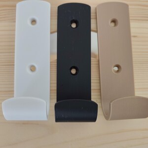 May include: Three wall-mounted hooks in white, black, and beige. Each hook features a vertical rectangular backplate with two circular screw holes for mounting and a curved, upward-facing hook at the bottom for hanging items securely.