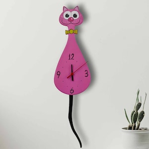 May include: Pink cat-shaped wall clock with a black tail and a yellow bow tie. The clock face has black numbers and a red hour hand.