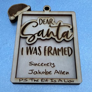 Könnte beinhalten: Ein Weihnachtsbaumschmuck aus Holz in Form eines Briefes mit dem Text "Lieber Weihnachtsmann, ich wurde hereingelegt Mit freundlichen Grüßen Jakobe Allen P.S. Der Elf ist ein Lügner"
