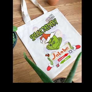 K&ouml;nnte beinhalten: Eine wei&szlig;e Canvas-Tragetasche mit einer gr&uuml;nen Grinch-Figur, die einen Weihnachtsmann-Hut tr&auml;gt. Die Tasche tr&auml;gt den Text "Special Delivery" und "To: Jakobe" mit einem H&auml;kchen neben "Nice" auf einer Liste von braven oder unartigen Kindern.