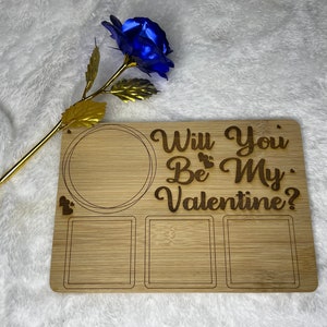 Könnte beinhalten: Ein hölzernes Valentinstags-Servierbrett mit dem Text "Willst du mein Valentin sein?" und einer blauen Metallrose mit goldenem Stiel. Das Brett hat einen kreisförmigen Bereich und zwei quadratische Bereiche.